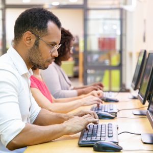 Extremely focused male student taking online test in computer class. Line of man and women in casual sitting at table, using desktops, typing, looking at monitor. Education center concept